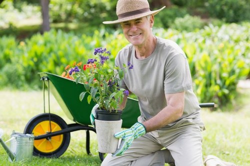 A gardener kneeling outdoors in a lush green garden, holding a potted flowering plant with purple, red, and orange blossoms. Behind them, a bright yellow and black wheelbarrow filled with assorted garden tools and a watering can rests on the grass, which has a dense, vibrant green appearance. In the background, there is a well-maintained hedge running along the edge of the garden, with some trees providing shade and creating a natural boundary. The scene is set on a clear day with natural daylight, highlighting the healthy foliage and freshly prepared garden area. The gardener wears a light grey T-shirt, a wide-brimmed straw hat, and gardening gloves, conveying professional outdoor maintenance work suitable for services such as hedge trimming and general gardening offered by Hedge Trimming Limehouse in the Limehouse area. The overall environment depicts a tidy and active outdoor space, reflecting the care and attention involved in horticultural tasks like planting, pruning, and garden landscaping.